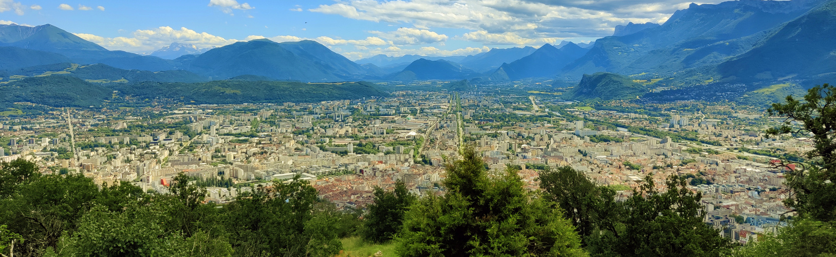 illustration-grenoble-panorama Vue panoramique de Grenoble depuis le mont Jalla avec des arbres verts en premier plan, puis la ville et les montagnes en arrière-plan